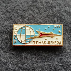 Puede incluir: Pin rectangular con borde dorado que presenta un globo terráqueo, un cohete estilizado y el texto "ЗЕМЛЯ-ВЕНЕРА" en blanco. El pin tiene un fondo azul y blanco con el año "1969" y las letras "CCCP".