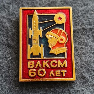 Puede incluir: Un pin rectangular de metal con fondo rojo, un cohete dorado y el perfil de una persona con casco espacial. Sobre el casco hay un pequeño círculo dorado con una estrella roja. El texto "ВЛКСМ 60 ЛЕТ" aparece en letras de color dorado.