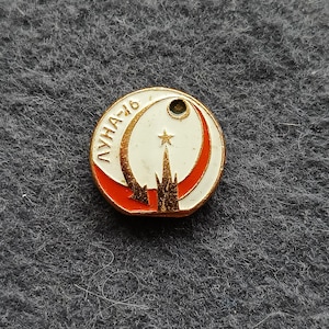 May include: A round, vintage pin with the text "ЛУНА-16" in gold lettering. The pin features a white background with a red crescent and a gold star. The bottom of the pin has a gold and red rocket design.