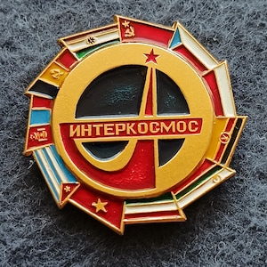 Puede incluir: Un pin metálico circular que presenta un emblema central de color dorado con el texto cirílico "ИНТЕРКОСМОС" en el medio. El diseño incluye una forma de cohete estilizada, estrellas rojas y un borde compuesto por varios segmentos de banderas nacionales.