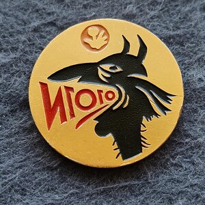 May include: A circular gold-colored metal pin featuring a black silhouette of a creature with a long snout and horns. The creature has its mouth open, revealing the red Cyrillic text "ИГОГО" inside. A small circular icon of a hand is at the top.