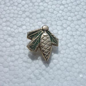 May include: A gold-colored metal pin in the shape of a pinecone with green leaf accents. The pin has a textured surface and is set against a white background.