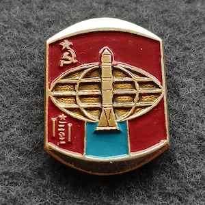 May include: A vintage pin with a gold border and a red background. The pin features a gold globe with a rocket, a hammer and sickle, and text. The bottom section is blue.