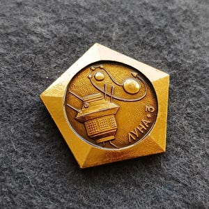 Puede incluir: Un pin vintage dorado con un marco pentagonal. El pin presenta un emblema circular con una nave espacial y el texto cirílico "ЛУНА 3". El diseño incluye una representación estilizada de un satélite y elementos celestes.