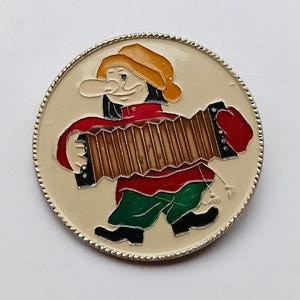 Può includere: Spilla rotonda color crema con l'immagine di un cartone animato di un musicista che suona la fisarmonica. Il musicista indossa una camicia rossa, pantaloni verdi e un cappello giallo. La spilla ha un bordo argentato e perlato.