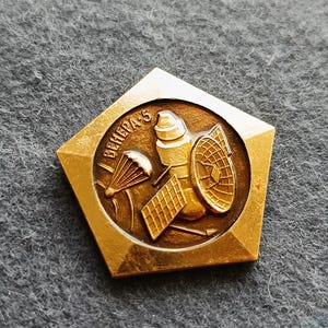 Puede incluir: Un pin dorado con forma pentagonal. El pin presenta un diseño circular central con un satélite, un paracaídas y un panel solar. El texto "ВЕНЕРА-5" está inscrito en la parte superior del círculo. El pin tiene un acabado metálico brillante.