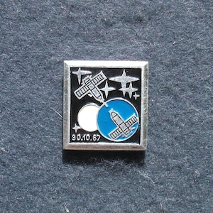 Puede incluir: Pin cuadrado plateado con fondo negro, diseño de satélite y estrellas blancas. Un círculo azul con un satélite está en la parte inferior derecha. La fecha "90.10.67" está en la parte inferior.