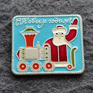 May include: A rectangular metal pin with a light blue background featuring a silver-toned train engine driven by a figure in a red and white suit. The text "С Новым годом!" is embossed in silver at the top. The border has a twisted rope design.