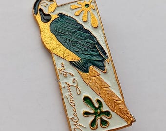 May include: A vintage rectangular pin featuring a stylized parrot design. The parrot has yellow and teal plumage, with gold accents and a white background. The pin includes text in a script font and decorative floral elements.