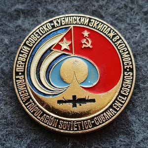 May include: A circular gold-toned metal pin featuring a red and blue enamel design with a central sun, a space station silhouette, and the Cuban flag. The border contains text in Russian and Spanish: Pervyy Sovetsko-Kubinskiy Ekipazh V Kosmose.