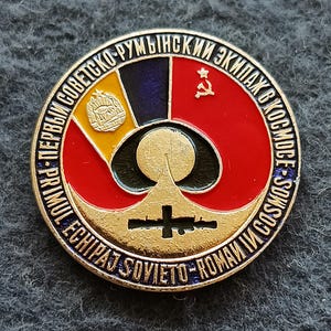 May include: A circular metal commemorative pin featuring a central gold rocket silhouette against a red, yellow, and blue background. It displays a hammer and sickle, a coat of arms, and text in Russian and Romanian: Pervyy Sovetsko-Rumynskiy Ekipazh V Kosmose.