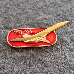 Pode incluir: Um alfinete vintage com um design de avião supersónico dourado, sobre um fundo vermelho. O alfinete tem o texto "VII 144" em dourado. O alfinete é retangular com bordas arredondadas.