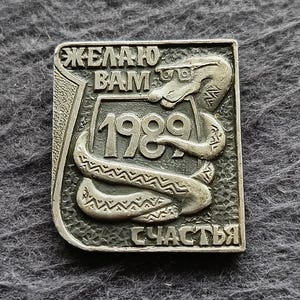 Könnte beinhalten: Eine rechteckige Anstecknadel aus silberfarbenem Metall mit einem Relief einer Schlange mit Brille, die sich um einen zentralen Schild windet. Der Schild zeigt das Jahr 1989. Kyrillischer Text lautet ЖЕЛАЮ BAM oben und СЧАСТЬЯ unten.