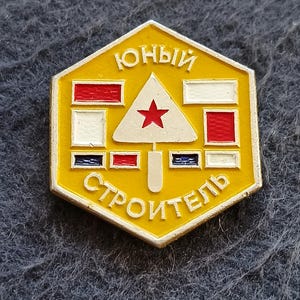Puede incluir: Un pin hexagonal de metal amarillo que presenta un triángulo blanco central con una estrella roja. El pin muestra el texto cirílico "ЮНЫЙ СТРОИТЕЛЬ" en letras blancas, junto con pequeños acentos rectangulares rojos, blancos y azules.
