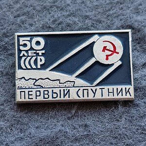 Puede incluir: Un pin rectangular de metal con fondo azul oscuro, un gráfico de satélite plateado y un símbolo rojo de martillo y hoz. El texto dice 50 ЛЕТ СССР y ПЕРВЫЙ СПУТНИК en letras blancas, e incluye una línea de horizonte estilizada en el diseño.
