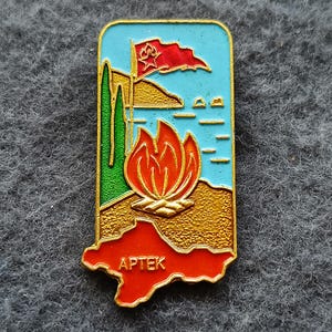 Puede incluir: Pin rectangular de esmalte con borde dorado. El pin presenta una bandera roja con una estrella, una llama estilizada y la palabra "APTEK" en blanco sobre un contorno de mapa rojo. El fondo incluye cielo azul y mar.