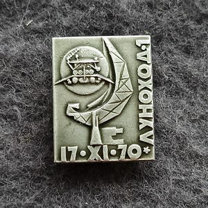 Puede incluir: Pin rectangular plateado con un diseño de vehículo lunar dentro de una media luna, con el texto "ЛУНОХОД" y la fecha "17·XI·70*". El pin tiene un diseño detallado en relieve.