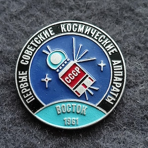 May include: A vintage round pin with a blue background featuring a satellite design and the text "CCCP". The pin has the text "PERBЫE COBETCKИE KOCMИЧECKИE AППAPATЫ" and "BOCTOK 1961" in Russian.