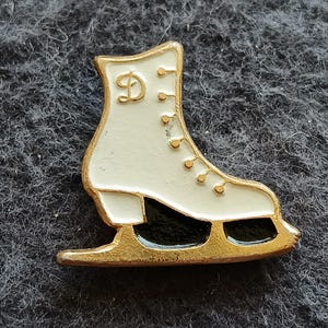 May include: A gold-toned metal pin shaped like an ice skate with a white boot, gold eyelets, and a black blade. The upper part of the boot features a gold cursive letter D. The design is outlined in gold metal, providing a clear contrast against the white enamel finish of the skate boot.