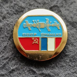 May include: A round, gold-colored pin featuring a blue background with a space station design and the text "CCCP FRANCE". The bottom section displays a red, white, and blue flag design with a hammer and sickle symbol.