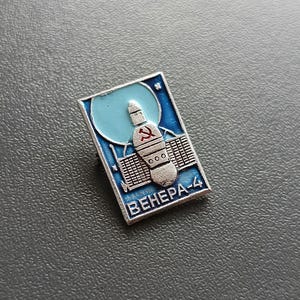 Puede incluir: Un pin de esmalte plateado y azul que representa una nave espacial con un símbolo de martillo y hoz rojo. El texto "ВЕНЕРА-4" está impreso debajo de la nave espacial.