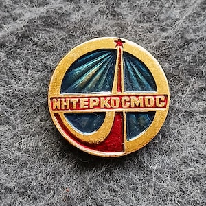 May include: A round, gold-colored pin with a blue and red design. The pin features the text "ИНТЕРКОСМОС" in red lettering. A red star is at the top of the design.
