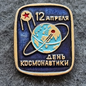 Puede incluir: Pin vintage con fondo azul y borde dorado. El pin presenta una imagen estilizada de la Tierra con un satélite y una estrella roja. También está presente el texto "12 Abril" y "Día de la Cosmonáutica".