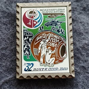 May include: A rectangular pin featuring a colorful design with the text "INTERKOSMOS" and Cyrillic text. The pin depicts a space capsule, astronauts, and a helicopter, with a stamp-like border. The number "32" and the text "ПОЧТА СССР. 1980" are also visible.
