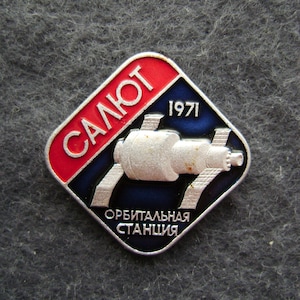 May include: A vintage metal pin with a space station design. The pin features the word "САЛЮТ" in red, the year "1971", and the words "ОРБИТАЛЬНАЯ СТАНЦИЯ" in white. The background is blue and the pin has a silver border.
