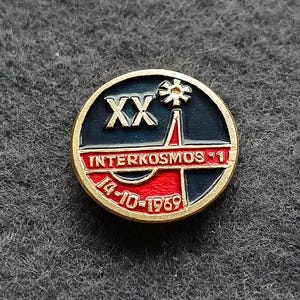 May include: A round, gold-toned pin with a dark blue and red design. The pin features the text "XX", "INTERKOSMOS 1", and "14-10-1969". A stylized image of a tower is also present. The pin has a gold border.