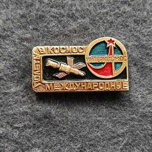 May include: A vintage gold-toned pin with the words "KOCMOC" and "MEЖДУНАРОДНЫЕ" in Cyrillic script. The pin features a satellite and a red star on a blue and red background, with the text "ИНТЕРКОСМОС".