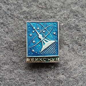 Puede incluir: Un pin cuadrado y vintage con un fondo de esmalte azul que presenta un diseño estilizado de lanzamiento de cohetes, estrellas y símbolos atómicos. El pin tiene la palabra "Байконур" en letras doradas en la parte inferior.
