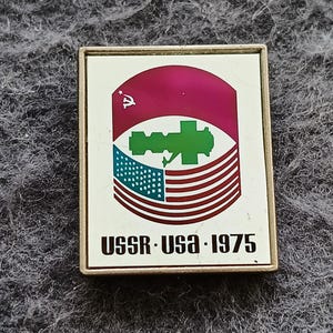 May include: A rectangular metal pin featuring a white background with a central graphic of a green space station silhouette between a red Soviet flag segment and a blue and white American flag. The text USSR-USA-1975 is printed in black.
