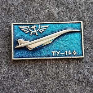 Pode incluir: Um alfinete retangular com fundo azul e design de avião prateado. O alfinete apresenta o texto "TY-144" e um símbolo de martelo e foice com asas. O alfinete tem uma borda prateada.