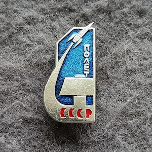 Puede incluir: Un pin vintage plateado con un fondo de esmalte azul. El pin presenta un cohete lanzándose al espacio, con la palabra "ПОЛЕТ" en letras cirílicas blancas. La parte inferior del pin tiene las letras "CCCP" en rojo.