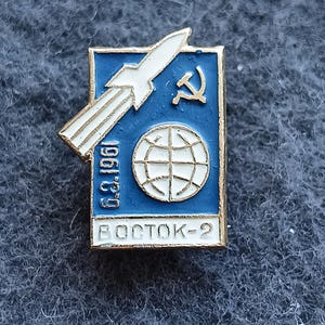 Puede incluir: Pin vintage de esmalte con un cohete blanco, una hoz y un martillo, y un globo terráqueo sobre fondo azul. El pin tiene el texto "1961 g." y "VOSTOK-2" en letras blancas, con un borde dorado.