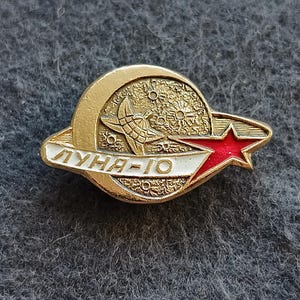 Puede incluir: Un pin vintage dorado con una luna creciente, estrellas y una estrella roja. El pin tiene el texto "ЛУНА-10" en letras blancas. Es probable que el pin sea un artículo conmemorativo.