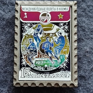 May include: A vintage postage stamp with a gray border. The stamp features a colorful illustration of astronauts in space, with text in Russian. The stamp has a value of 15 and the text "ПОЧТА СССР".