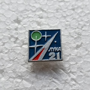 Puede incluir: Pin cuadrado con fondo azul, con un círculo verde, una cruz blanca y un diseño rojo y plateado. El texto "ЛУНА 21" está impreso en plata. Un objeto de colección vintage de exploración espacial.
