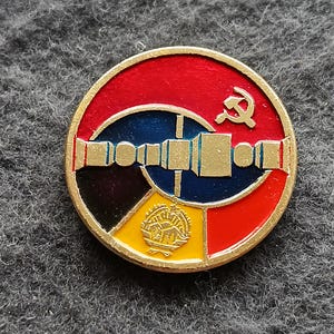 May include: A round, enameled pin with a gold border. The pin features a red top section with a hammer and sickle, a blue and gold center, and a yellow and black bottom section. The pin is likely a vintage collectible.