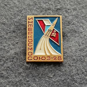 May include: Rectangular vintage pin with a rocket design. The pin features a blue background with a red and white V-shaped element. A rocket is depicted launching, with the text "СОЮЗ 3.28" and other Cyrillic text.