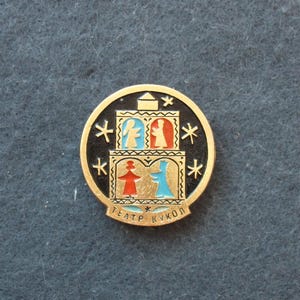 Könnte beinhalten: Runder, goldfarbener Ansteckpin mit schwarzem Zentrum. Er zeigt eine stilisierte Illustration eines Gebäudes mit Figuren in Rot, Blau und Weiß. Der Pin trägt den weißen Text "TEATP KYKOЛ".