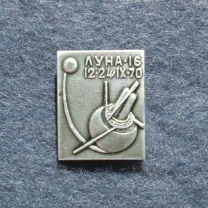 Puede incluir: Pin rectangular plateado con el texto "ЛУНА-16 12-24 IX-70". El pin tiene un diseño estilizado de una esfera con líneas y un círculo. El pin está sobre una superficie azul oscuro texturizada.