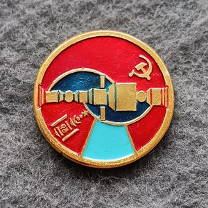 May include: A round, red and gold pin with a blue and gold satellite design. The pin features a hammer and sickle symbol and text in a foreign language. The pin is likely a vintage collectible.