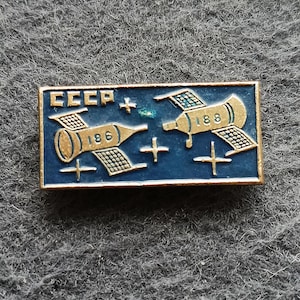 Puede incluir: Un pin rectangular vintage con fondo azul y detalles dorados. El pin presenta dos naves espaciales estilizadas con los números "186" y "188", y las letras "CCCP". Tiene un borde dorado y símbolos de cruz.