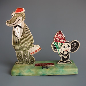 May include: A metal figurine set featuring a crocodile in a pinstripe suit and red hat holding a cake, and a small creature with large ears holding red flowers. A plaque on the green marble-effect base reads "С ДНЕМ РОЖДЕНИЯ!"