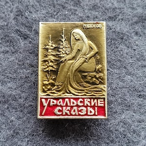 May include: A rectangular gold-colored pin with a relief of a woman seated near a pine tree. The pin has the text "УРАЛЬСКИЕ СКАЗЫ" in red letters. The text "ПЕАЖОВ" is at the top of the pin.