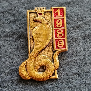 May include: A gold-toned metal pin featuring a textured cobra wearing a small crown. To the right of the snake, a vertical rectangular panel contains the red-filled numbers 1, 9, 8, and 9, each stacked within its own individual square section.