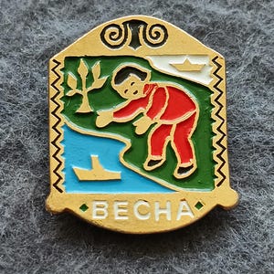 May include: A gold-colored pin with a colorful design featuring a person in red clothing reaching towards a body of water. The pin has a green and blue landscape with a tree and boats. The word "BECHA" is at the bottom.