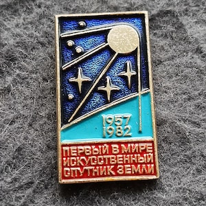 Puede incluir: Pin rectangular con fondo azul y turquesa, con un diseño de satélite y los años 1957 y 1982. El pin tiene un borde dorado y texto en rojo.
